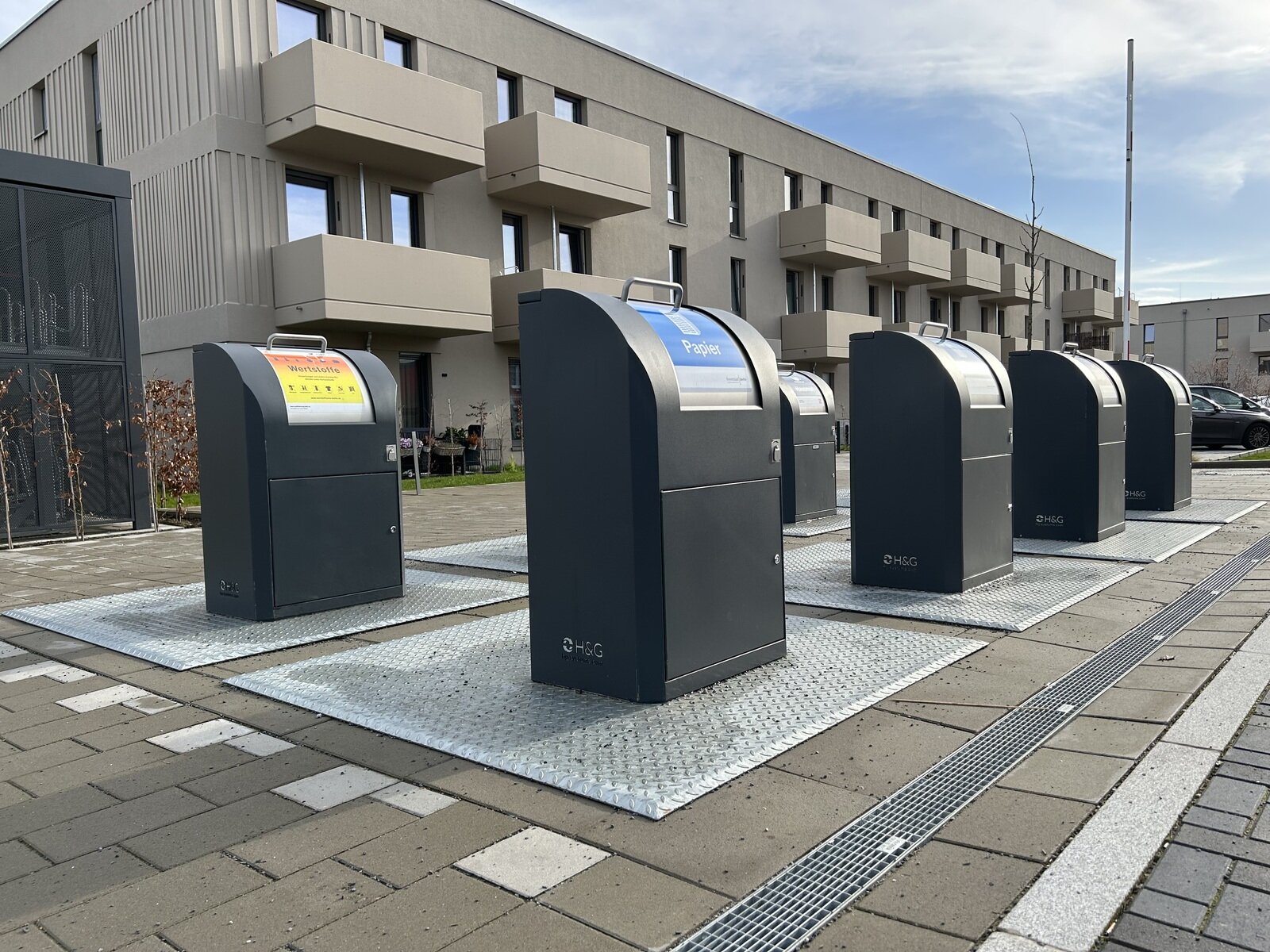 Sechs Unterflurbehälter in einer Reihe auf befestigten Plattformen vor einem mehrgeschossigen Wohnhaus. Von jedem Behälter ist nur die etwa hüfthohe Einwurfsäule zu sehen.
